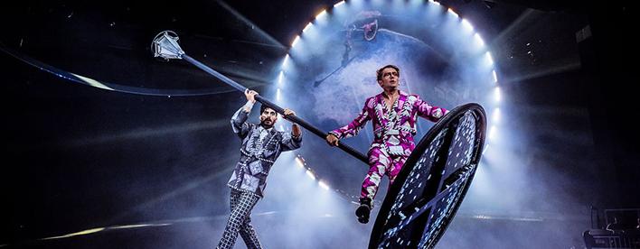 Two performers in colorful patterned suits on a dramatic stage, one holding a long pole attached to a large circular prop, with a glowing ring of light and stage fog in the background.