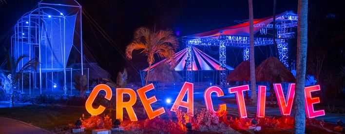 Illuminated outdoor setup at night featuring a large neon “CREACTIVE” sign in red and orange tones. Behind it are circus-style structures lit in blue, including rigging towers, a tent-like canopy, and palm trees.
