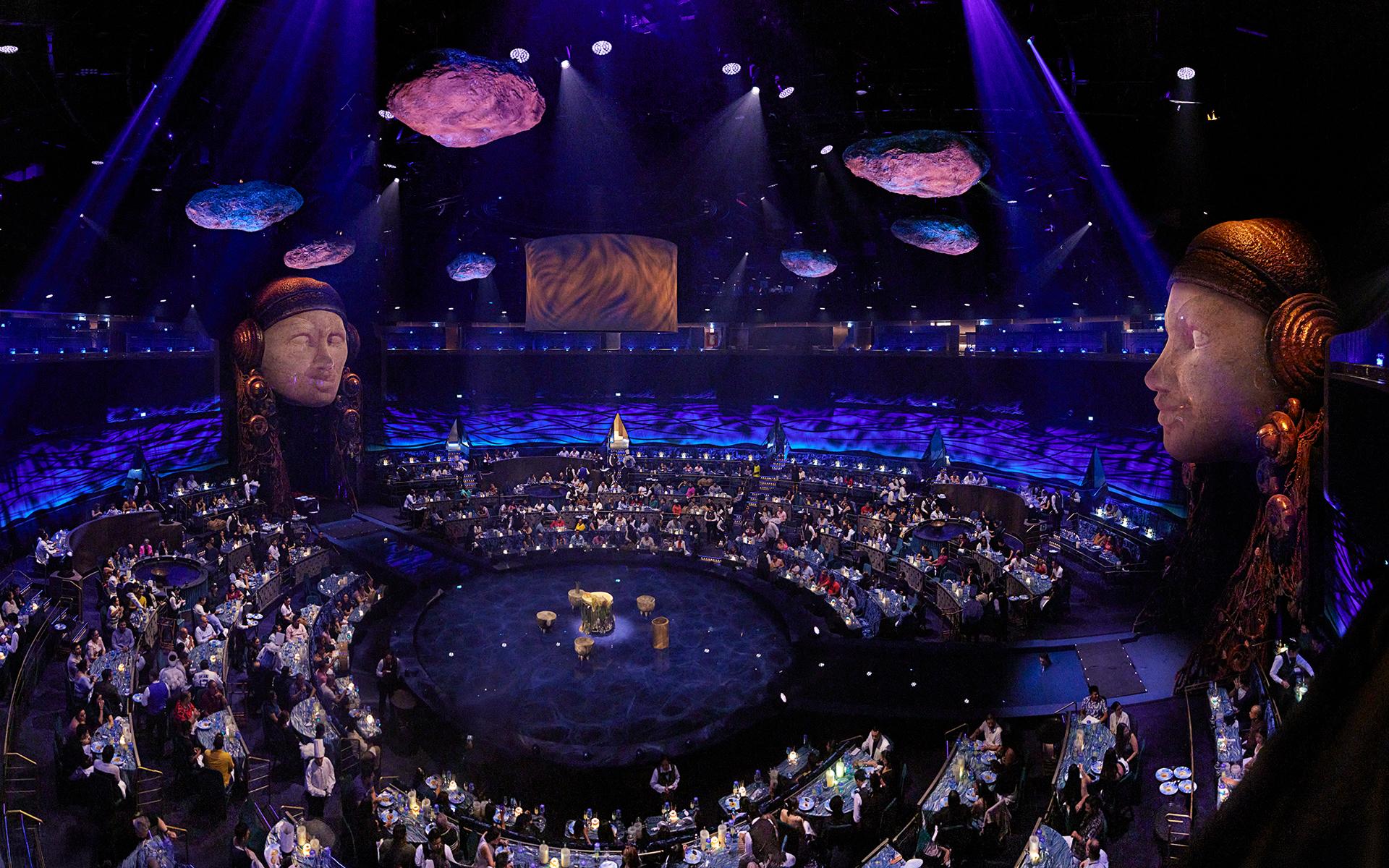 Panoramic view of a large immersive dinner theater with guests seated around a circular stage, dramatic purple lighting, floating rock-like set pieces, and monumental sculpted heads overlooking the audience.