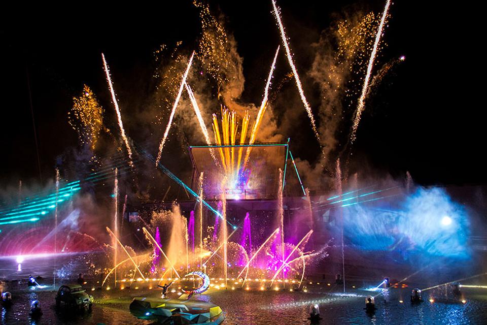 Cirque du Soleil water stage illuminated with fireworks, fountains, and colorful light effects during a spectacular nighttime performance.