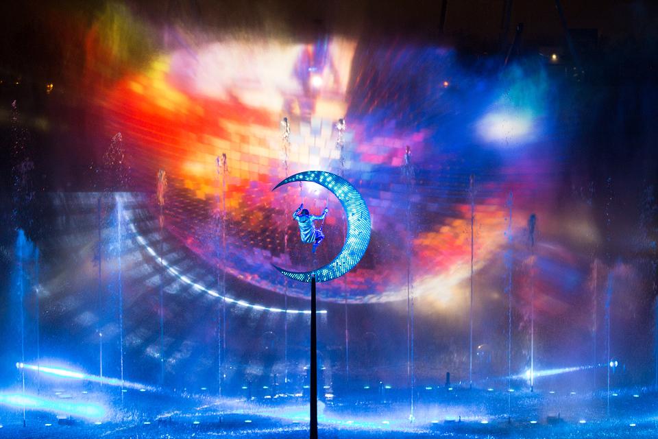 Aerial performer on a glowing crescent moon above a water stage, surrounded by swirling multicolored projections and blue lighting.