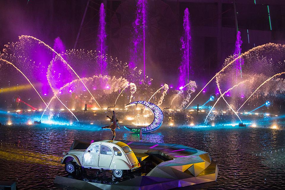 Aerial performer on a glowing crescent moon above a water stage, surrounded by swirling multicolored projections and blue lighting.