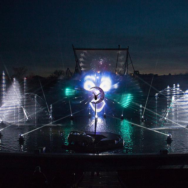 Wide view of Cirque du Soleil’s aquatic stage with synchronized fountains, crescent moon set piece, and immersive lighting effects at night.