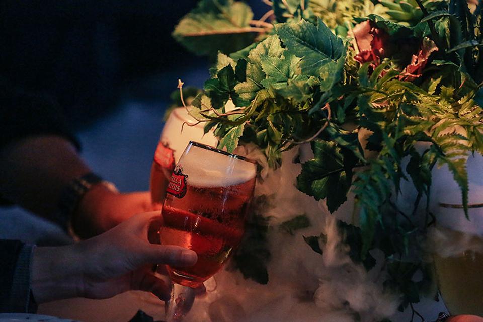 Guest holds a Stella Artois beer beside a lush floral centerpiece with mist effects at a Cirque du Soleil experiential dining event.