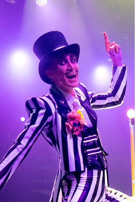 Cirque du Soleil performer in a black-and-white striped costume and top hat smiles and gestures playfully under colorful stage lighting.