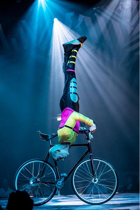 Cirque du Soleil acrobat performs a handstand on a bicycle under dramatic blue stage lighting.