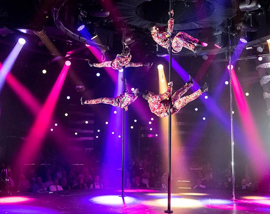 Cirque du Soleil aerial artists perform synchronized pole acrobatics high above the stage with colorful spotlights and a live audience below.