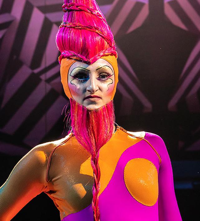Close-up portrait of a Cirque du Soleil performer in vibrant pink and orange costume with bold theatrical makeup and sculptural hairstyle.