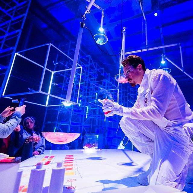 Cirque du Soleil performer in a white costume prepares a theatrical experiment on an interactive stage surrounded by guests under blue lighting.