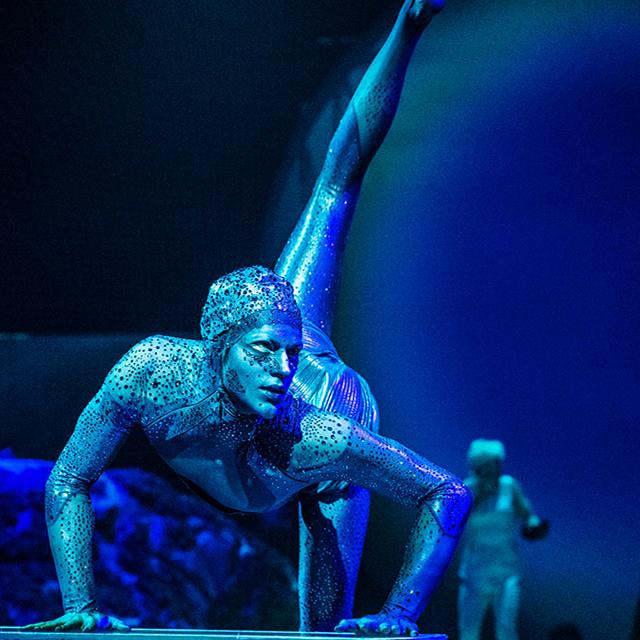 Cirque du Soleil contortionist in a shimmering blue costume performs a dramatic floor pose under spotlight on stage.