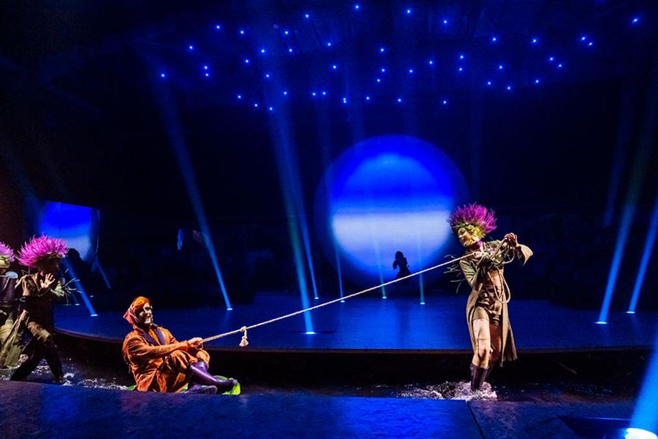Cirque du Soleil performers act out a playful scene on a water stage using ropes and props beneath a starry blue lighting design.