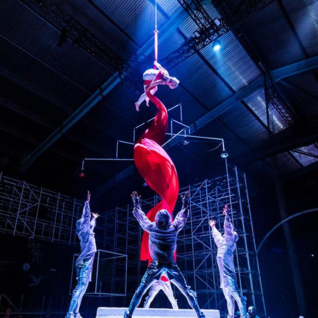 Cirque du Soleil aerial performers lift and spin a red silk acrobat high above the stage inside a large theatrical venue.