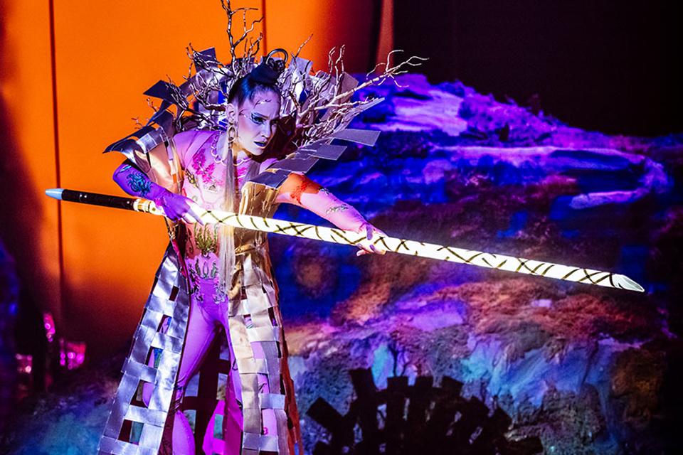 Cirque du Soleil aerial performers lift and spin a red silk acrobat high above the stage inside a large theatrical venue.