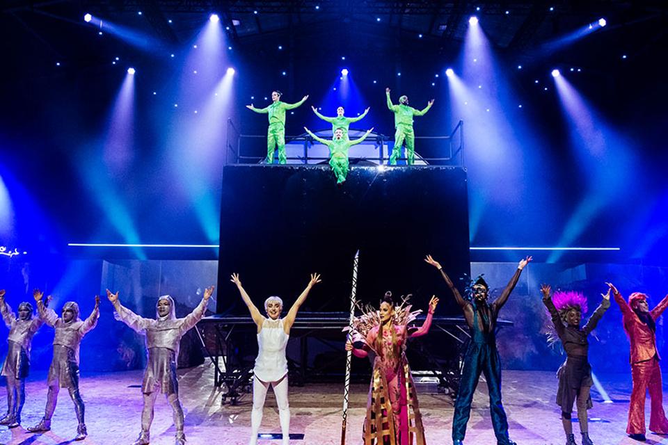 Cirque du Soleil cast takes a curtain call on stage, raising their arms under bright blue and purple spotlights.