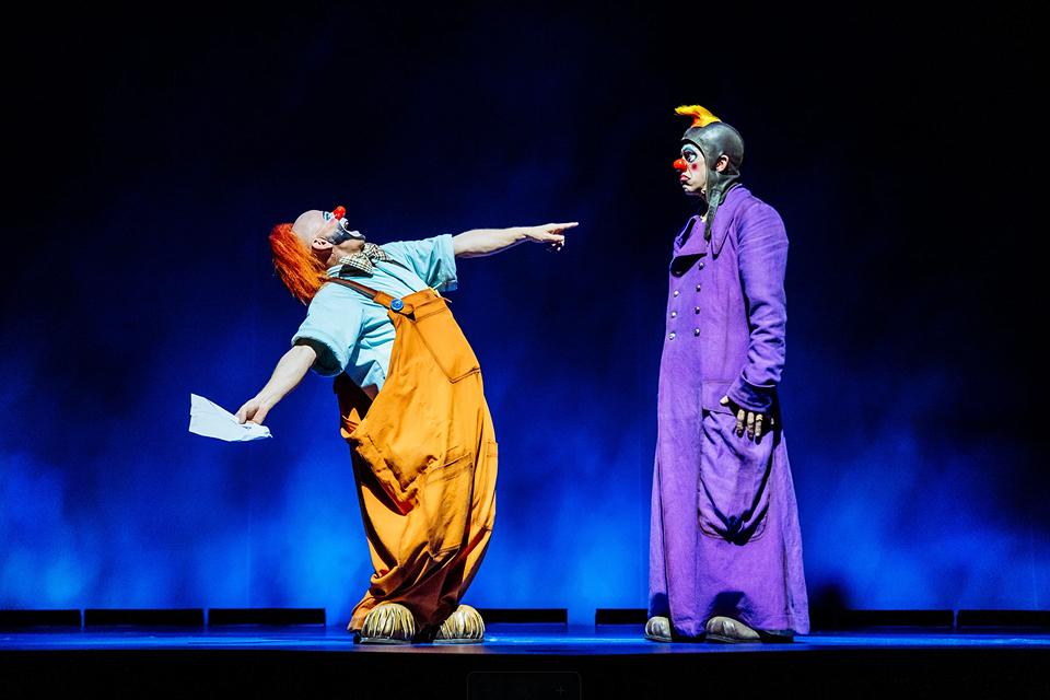 Two Cirque du Soleil clown characters perform a playful sketch on stage, wearing colorful costumes under blue theatrical lighting.