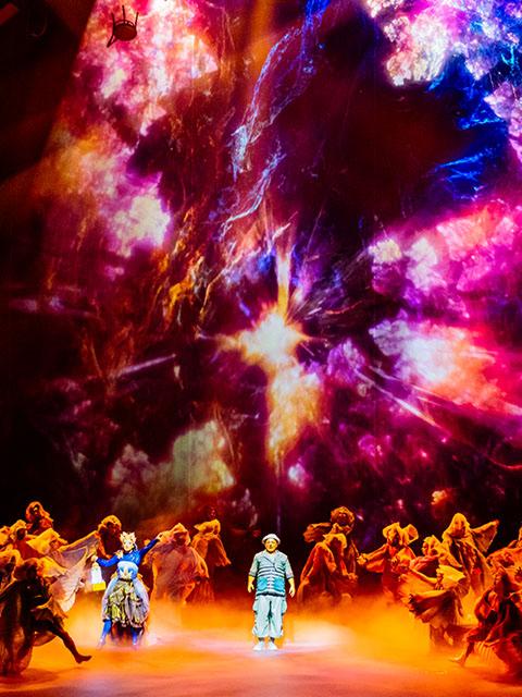 Two Cirque du Soleil clown characters perform a playful sketch on stage, wearing colorful costumes under blue theatrical lighting.