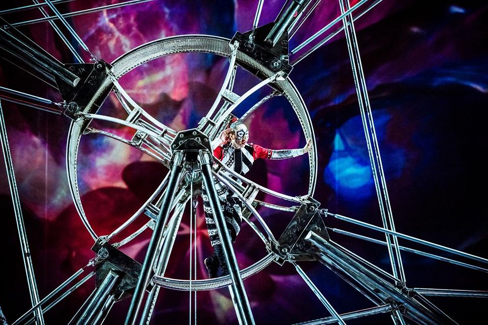 Cirque du Soleil acrobat performs inside a large rotating metal wheel against a vivid, abstract digital background.