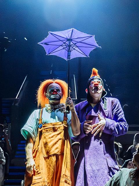 Two Cirque du Soleil clowns stand on stage holding an umbrella, wearing colorful costumes under soft blue theatrical lighting.