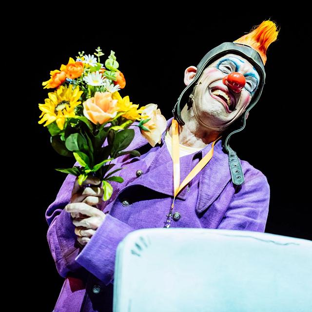 Cirque du Soleil clown in a purple costume smiles while holding a bouquet of yellow and orange flowers on stage.