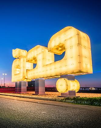 Illuminated “Jeddah” landmark sign glowing at dusk along a roadside under a clear blue sky.