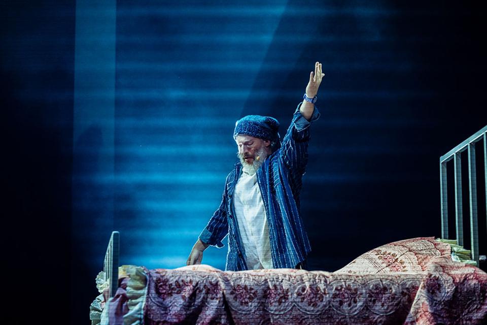 Cirque du Soleil performer in striped pajamas raises his hand while standing on a bed under blue stage lighting.