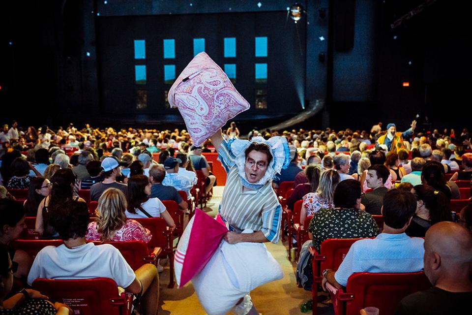 Cirque du Soleil character carries oversized pillows through a seated audience, interacting playfully with spectators.
