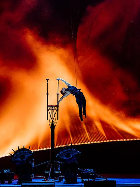 Cirque du Soleil acrobat performs a high jump and pole routine against a fiery orange and red stage backdrop.