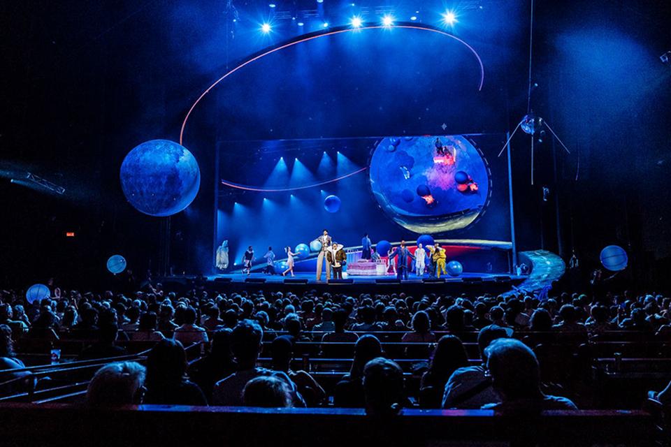 Wide view of a Cirque du Soleil arena show featuring aerial apparatus, floating spheres, and a packed audience under blue lighting.