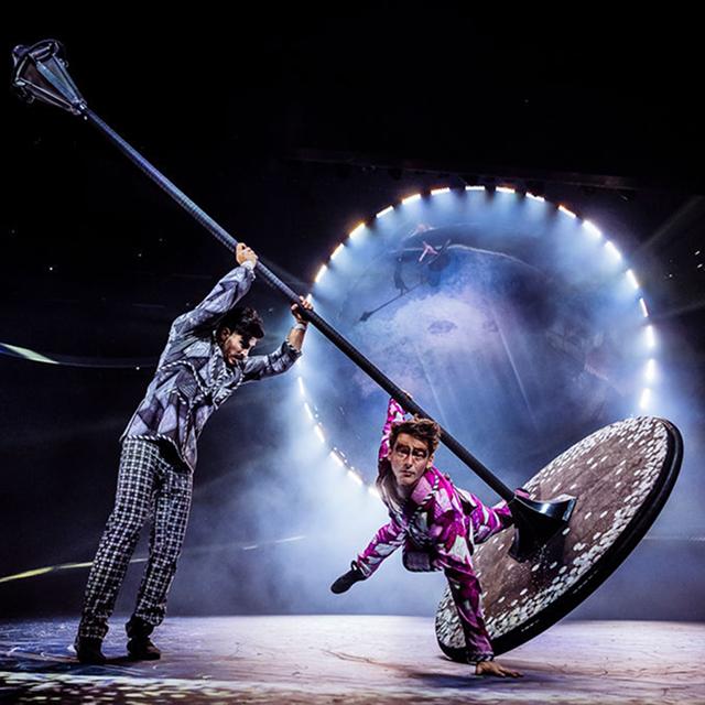 Cirque du Soleil performers balance and spin on a large rotating pole and disc apparatus under dramatic stage lighting.