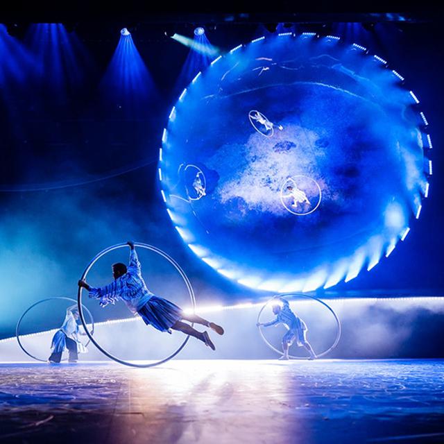 Cirque du Soleil acrobats perform Cyr wheel routines in front of a glowing circular backdrop on a blue-lit stage.
